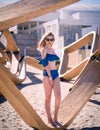Blonde Caucasian Woman on the Beach With Blue Flounce Top Swimsuit, Sitting on a Wood Structure and Looking Away