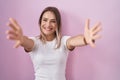 Blonde caucasian woman standing over pink background looking at the camera smiling with open arms for hug Royalty Free Stock Photo