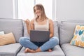 Blonde caucasian woman using laptop at home sitting on the sofa pointing thumb up to the side smiling happy with open mouth Royalty Free Stock Photo