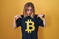 Blonde caucasian woman wearing bitcoin t shirt pointing down with fingers showing advertisement, surprised face and open mouth