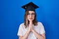 Blonde caucasian woman wearing graduation cap praying with hands together asking for forgiveness smiling confident