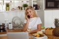 Blonde cheerful young woman talking on cellphone at laptop in kitchen with white wine. Royalty Free Stock Photo