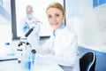 Blonde chemist in white coat holding eyeglasses and working with microscope