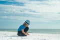 Blonde child in a hat and shirt draw pictures in the sand by the sea. Travel and recreation with children. Childrens