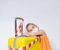 Blonde child in orange blouse, medical mask. Posing isolated on white. She leaning yellow suitcase tied white and red