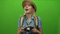 Blonde explorer woman in straw hat raising camera with both hands and gripping backpack strap in studio; curiosity Royalty Free Stock Photo
