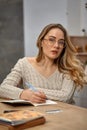 Blonde female blogger in glasses, beige sweater. Sitting in kitchen at wooden table, going to write something in her Royalty Free Stock Photo