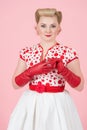 Blonde female pin-up styled pretty woman removes the red glove from the hand isolated in studio with pastel pink background