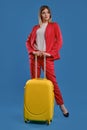 Blonde female in red pantsuit, white blouse and high black heels. She is holding yellow suitcase, posing against blue Royalty Free Stock Photo