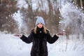 blonde girl in a black fur coat and a blue hat against the backdrop of a winter snowy forest. She throws snow and it flies in the Royalty Free Stock Photo