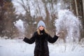 blonde girl in a black fur coat and a blue hat against the backdrop of a winter snowy forest. She throws snow and it flies in the Royalty Free Stock Photo