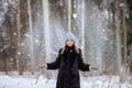 blonde girl in a black fur coat and a blue hat against the backdrop of a winter snowy forest. She throws snow and it flies in the Royalty Free Stock Photo