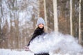 blonde girl in a black fur coat and a blue hat against the backdrop of a winter snowy forest. She throws snow and it flies in the Royalty Free Stock Photo