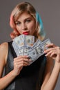 Blonde girl in black stylish dress holding some money, posing against gray background. Gambling entertainment, poker