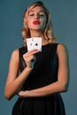 Blonde girl in black stylish dress showing two playing cards, posing against blue background. Gambling entertainment Royalty Free Stock Photo