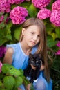 A blonde girl in a blue dress holds a black cat with white spots in her arms, a girl in the garden