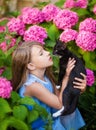 A blonde girl in a blue dress holds a black cat with white spots in her arms, a girl in the garden