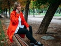 Blonde girl with blue eyes and dressed in red gabardine talks on the phone going up to a bench in a park