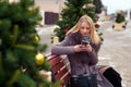 Blonde girl in a brown coat with fur sits on a wooden bench in the park and reads sms on a mobile phone. near christmas trees Royalty Free Stock Photo