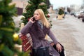 Blonde girl in a brown coat with fur sits on a wooden park bench and talking on a cell phone. near christmas trees Royalty Free Stock Photo