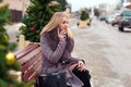 Blonde girl in a brown coat with fur sits on a wooden park bench and talking on a cell phone. near christmas trees Royalty Free Stock Photo