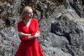 A blonde girl with fluttering hair in the wind in a red dress in an elegant manner stands on the bank of a mountain river with Royalty Free Stock Photo