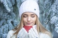 Blonde girl in a frozen conifer forest smells hot drink in a red cup with eyes closed, all dressed in white winter clothes Royalty Free Stock Photo