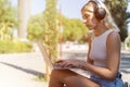 A blonde girl with headphones sitting on a bench on a sunny day, using a laptop. Royalty Free Stock Photo