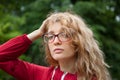 Blonde girl with hipster eyeglasses in dark red jacket looking at the camera posing outdoors in the urban park