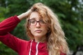 Blonde girl with hipster eyeglasses in dark red jacket looking at the camera posing outdoors in the urban park