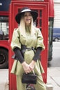 A blonde girl with long hair in a green pantsuit and wide-brimmed hat poses in front of a red telephone box