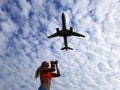 Blonde girl with long hair takes pictures of a plane flying over her on a cloudy sky background