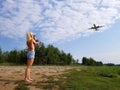 Blonde girl with long hair takes pictures of a plane flying over her on a cloudy sky background