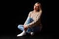 A blonde girl sits on the floor in the studio on a black background in jeans, a sweater and socks, her legs are crossed, she looks Royalty Free Stock Photo