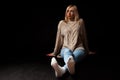 A blonde girl sits on the floor in the studio on a black background in jeans, a sweater and socks, her legs are stretched and Royalty Free Stock Photo