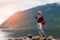 A blonde girl in sportswear with a red backpack on her shoulder and phone stands on the stones against the backdrop of a mountain