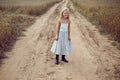 Blonde girl stands on an earthen path in a long dress among a wheat field