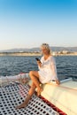 A blonde girl with sunglasses on her head holds a smartphone in her hands, traveling the sea on a yacht. White pareo on
