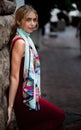 Blonde girl wearing a maroon dress and blue scarf leaning against a stone wall in an urban setting Royalty Free Stock Photo