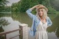 The blonde holds a straw hat on her hand and stands on the edge of a stone pier in a white dress and a blue denim shirt