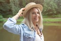 The blonde holds a straw hat on her hand and stands on the edge of a stone pier in a white dress and a blue denim shirt