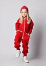 Blonde kid girl in white sneakers, red coldproof jumpsuit and winter hat stands lifting foot to step over, walking