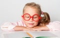 Blonde kid in red framed glasses, pink blouse. Smiling, leaning on table with copybook and pencil on it. Posing isolated