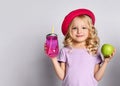 Blonde kid in red hat and purple blouse. She smiling, holding pink cocktail bottle and green apple, posing isolated on white Royalty Free Stock Photo