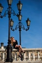 Blonde lady in black dress sitting on baluster railing under vintage street lamp Royalty Free Stock Photo