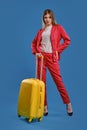 Blonde lady in red pantsuit, white blouse and high black heels. She is holding yellow suitcase, posing against blue Royalty Free Stock Photo