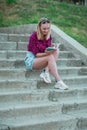 Blonde in light blue high-rise shorts, red plaid shirt, sneakers sitting on the stairs outside, read books. vertical photography