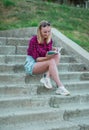 Blonde in light blue high-rise shorts, red plaid shirt, sneakers sitting on the stairs outside, read books. vertical photography