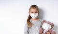 Little model in gray jumper and medical mask, posing isolated on white. Holding toy teddy bear which is also in mask