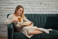 Blonde model blogger in beige knitted dress and socks is reclining on gray sofa, holding brown cup and reading book Royalty Free Stock Photo